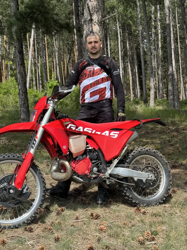 Mihail Todorov wearing black and red motocross gear standing behind a red GasGas enduro bike in a forest clearing.