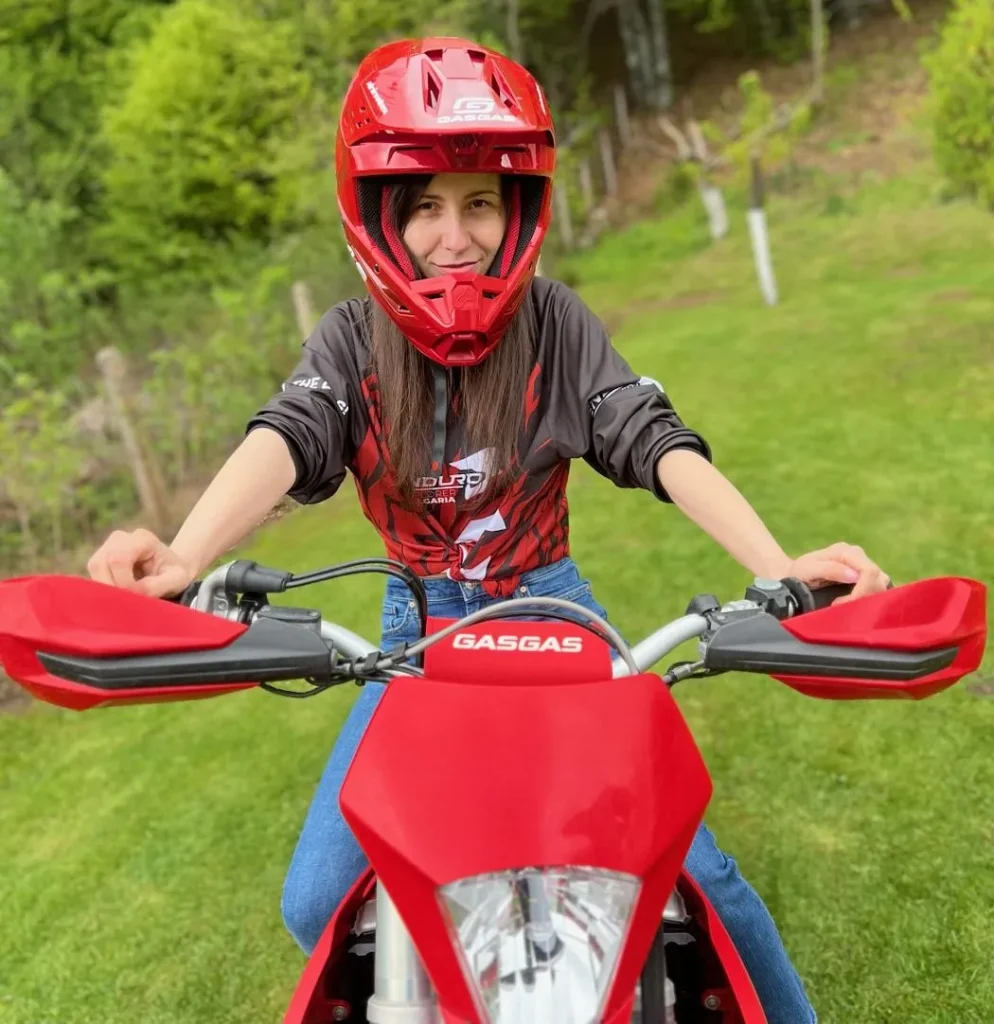 Stanislava Dimitrova wearing a red helmet and dark jacket sitting on a red GasGas enduro bike on green grass.