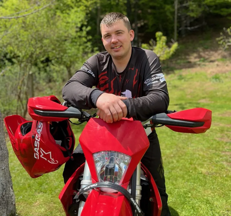Smiling Martin Spasov
leaning on the handlebars of a red enduro motorcycle outdoors with a red helmet hanging on the side.