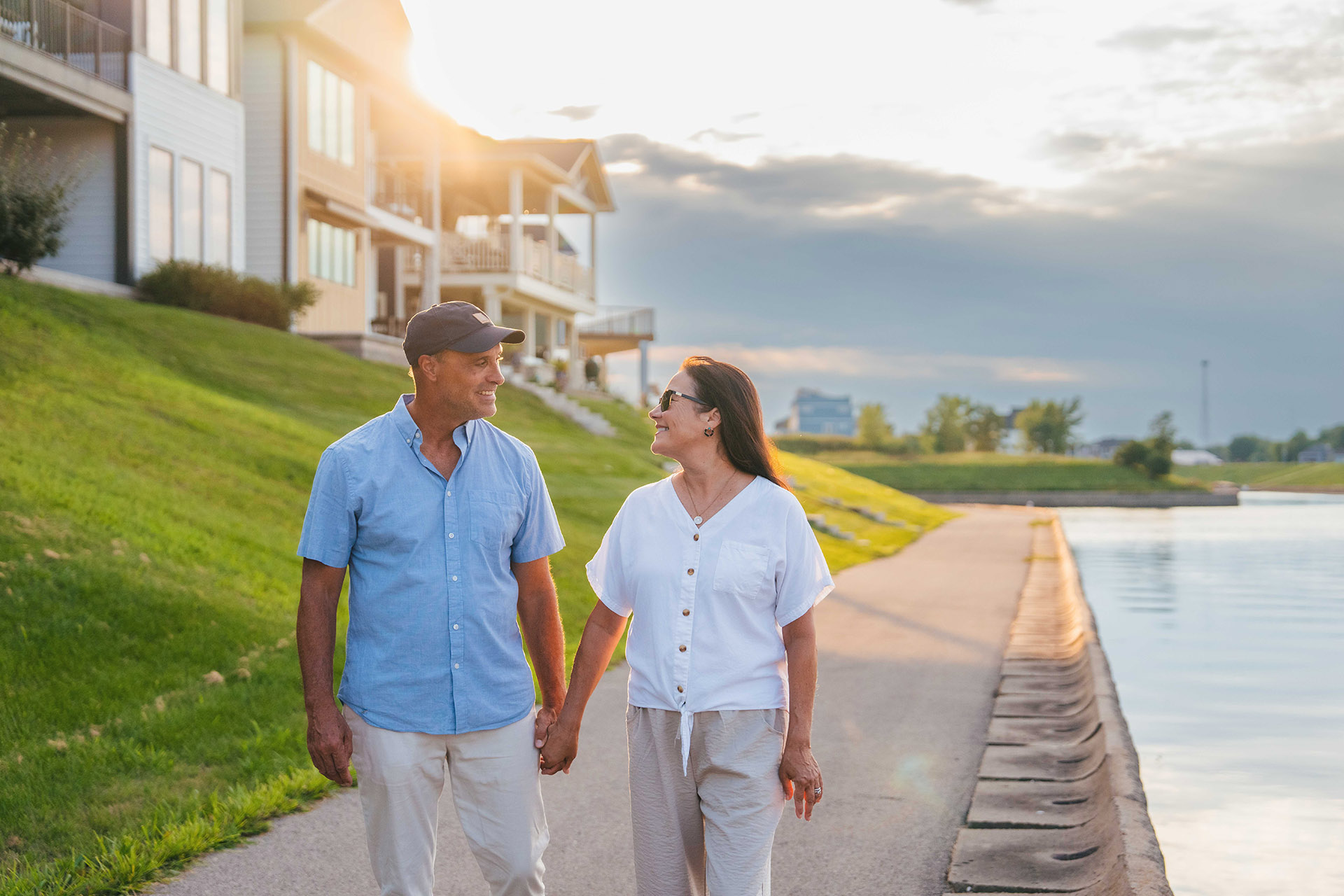 Middle-aged couple holding hands and walking along a waterfront pathway at sunset.