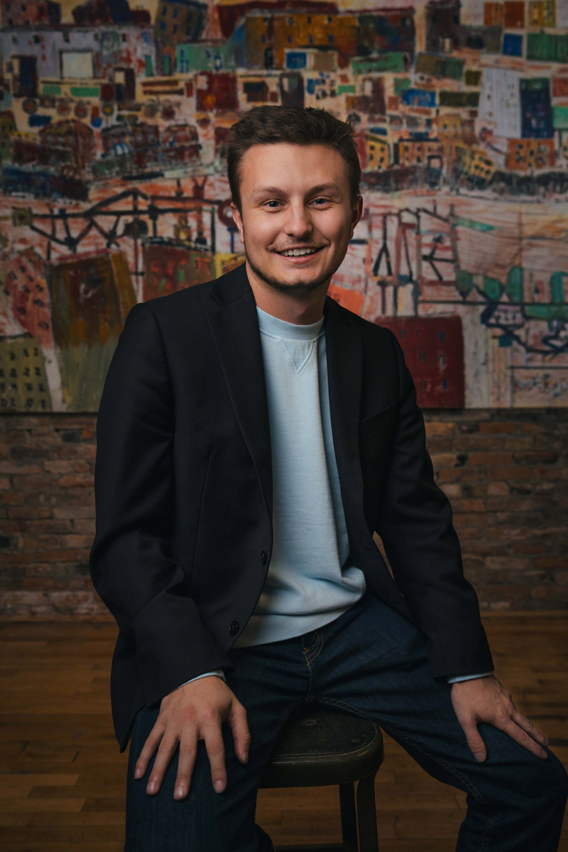 Matthew in a black blazer and light blue shirt sitting on a stool against a colorful mural background.