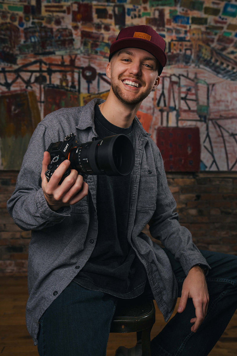 Kyle wearing a burgundy cap and grey shirt sitting on a stool and holding a camera with a large lens.