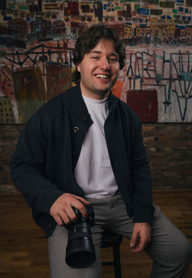 Connor sitting on a stool holding a camera, with a colorful abstract painting and brick wall in the background.