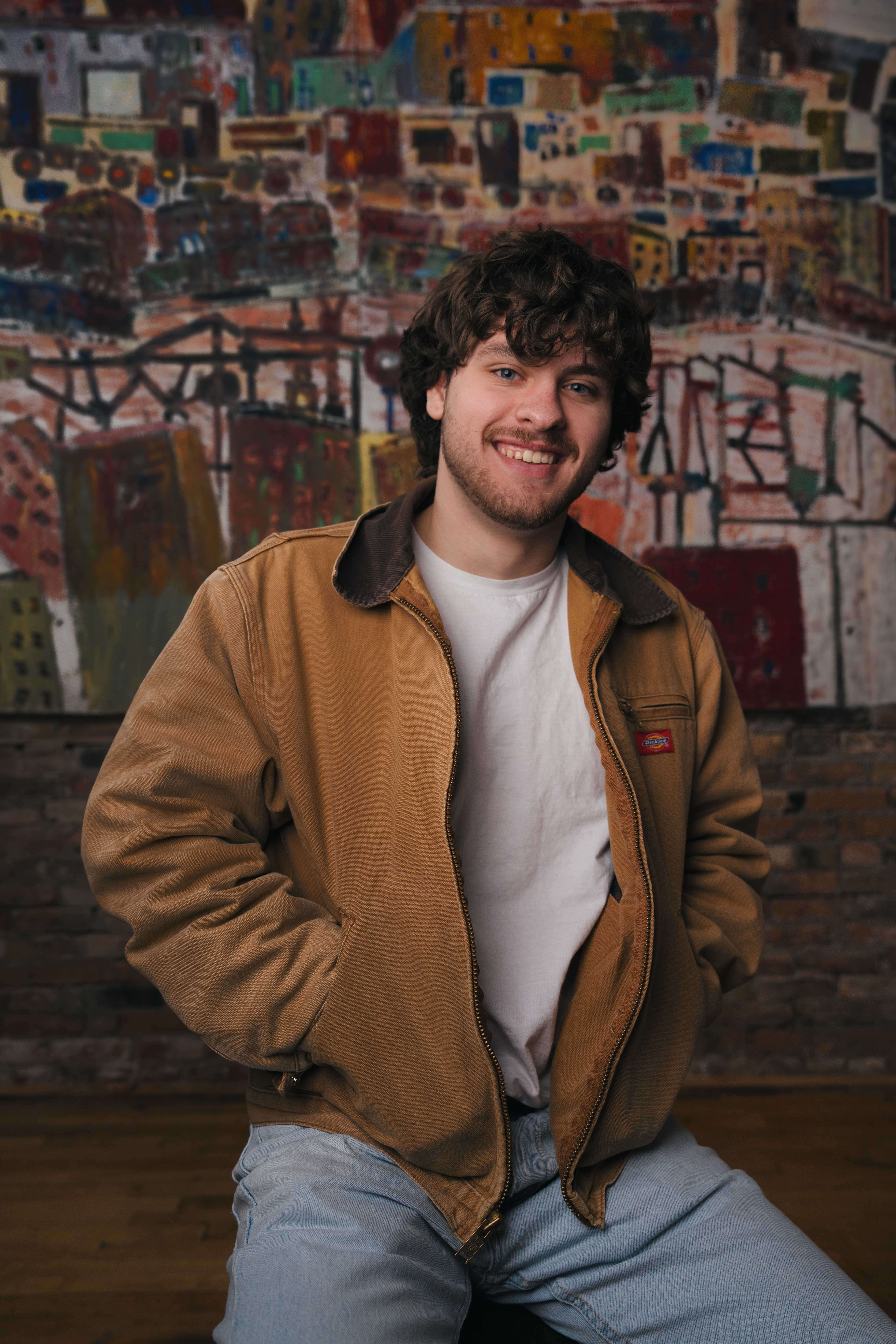 Smiling young man with curly hair wearing a brown jacket and white shirt sitting indoors.