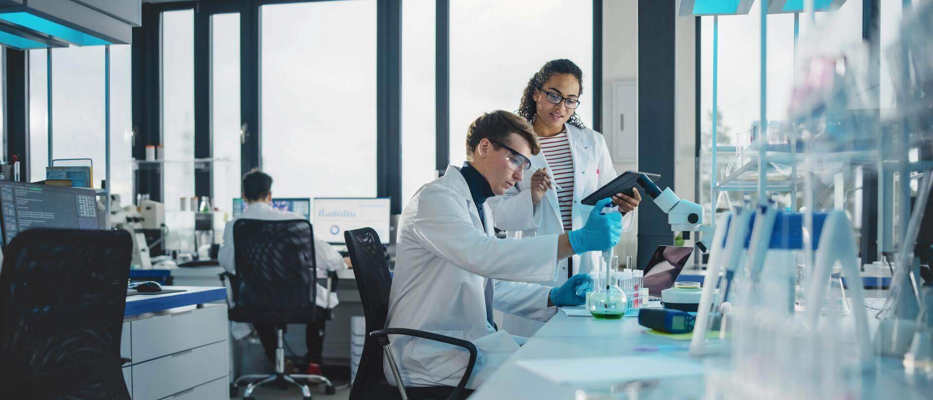 Two scientists in white coats work in a laboratory, one sitting and using a pipette while the other holds a digital table.