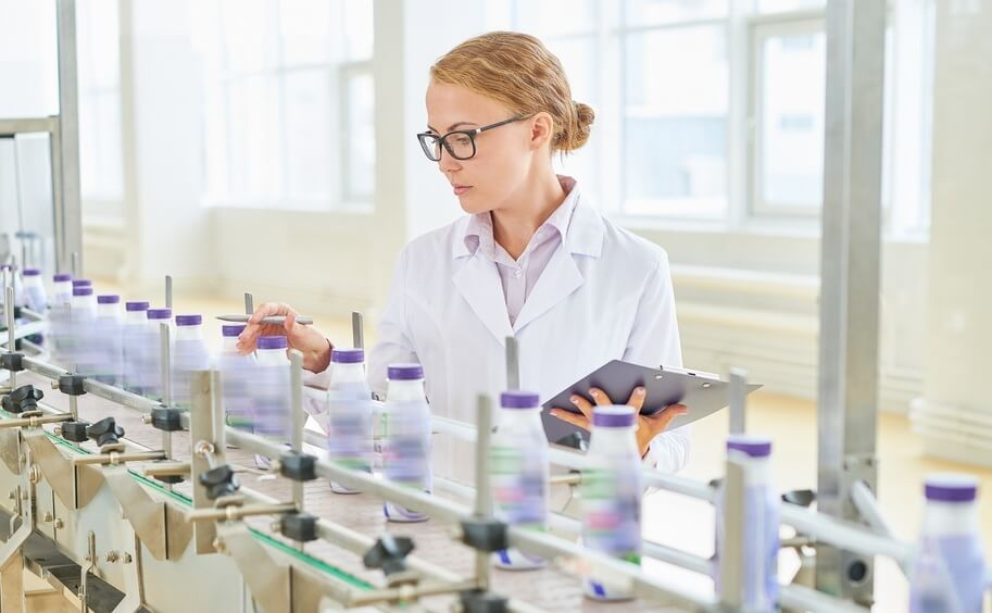 Frau im weißen Laborkittel prüft abgefüllte Flaschen auf einem Förderband in einer hellen Produktionsanlage.