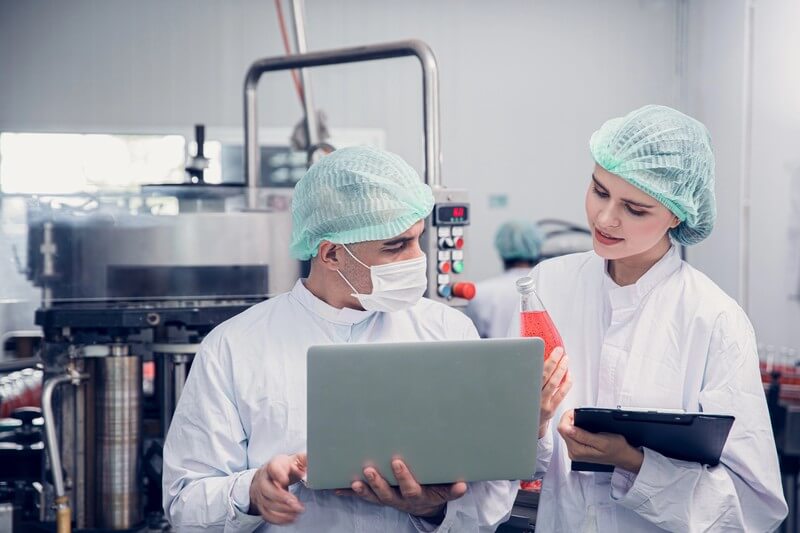 Zwei Mitarbeiter in weißer Schutzkleidung, Haarnetz und Maske überprüfen eine rote Getränkeflasche in einer Lebensmittelproduktionsanlage.