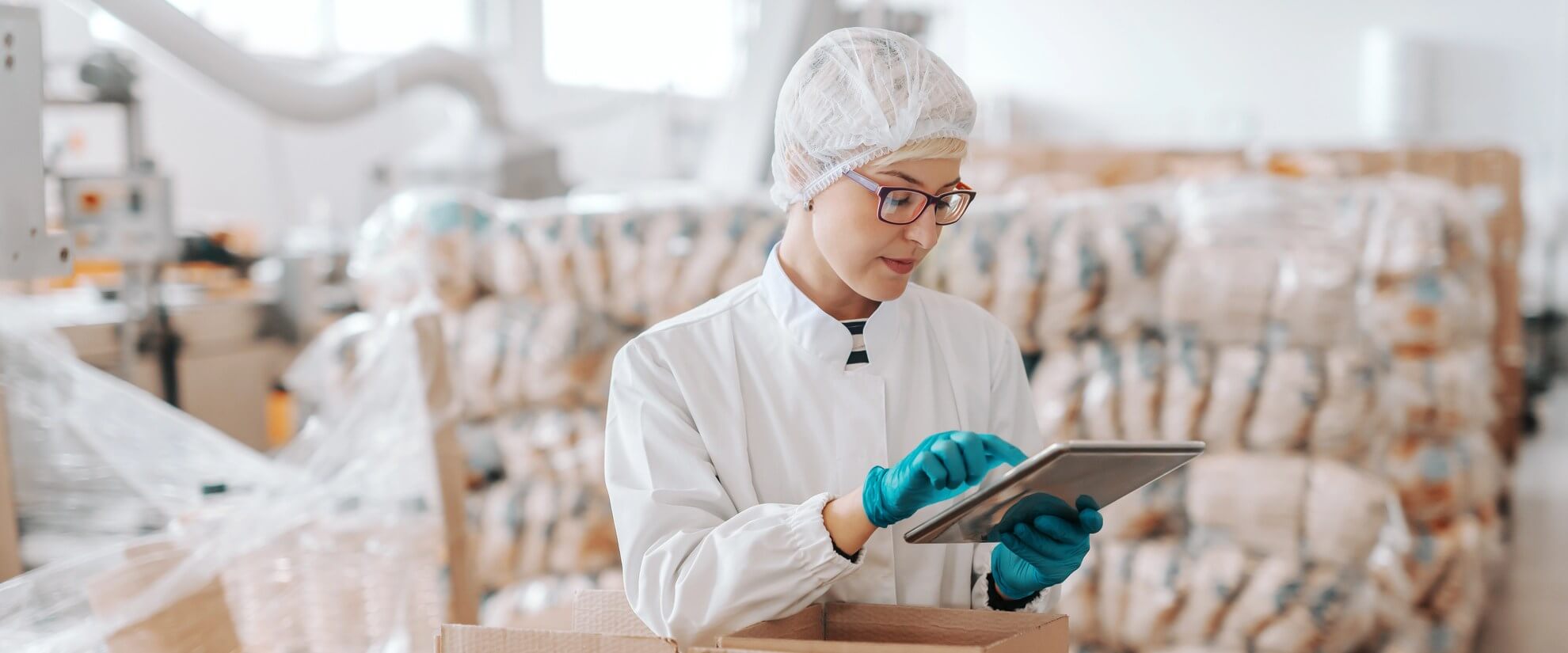 Frau in weißem Laborkittel, Haarnetz und blauen Handschuhen arbeitet mit Tablet in einer Lebensmittelverpackungsanlage.