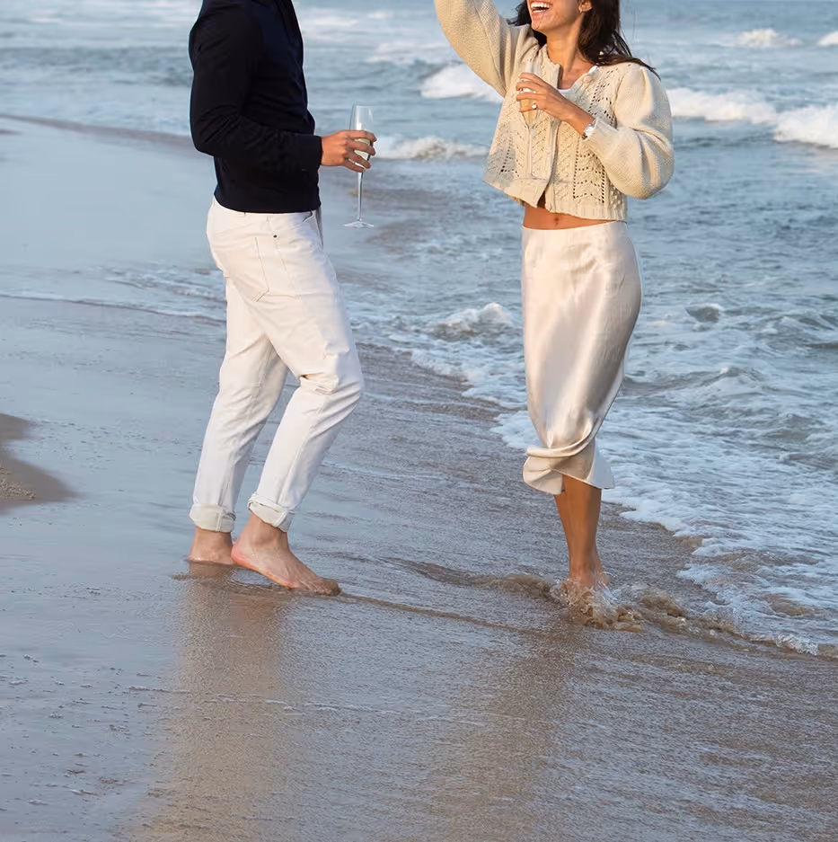 A man and woman standing barefoot and smiling on a beach near the ocean waves, each holding a glass of champagne.