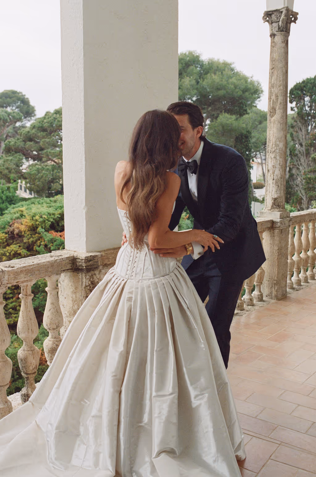Bride in a white corset wedding gown and groom in a black tuxedo embracing on a balcony with stone balustrades and green trees in the background.