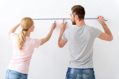 Man and woman measuring distance on a white wall using a tape measure.