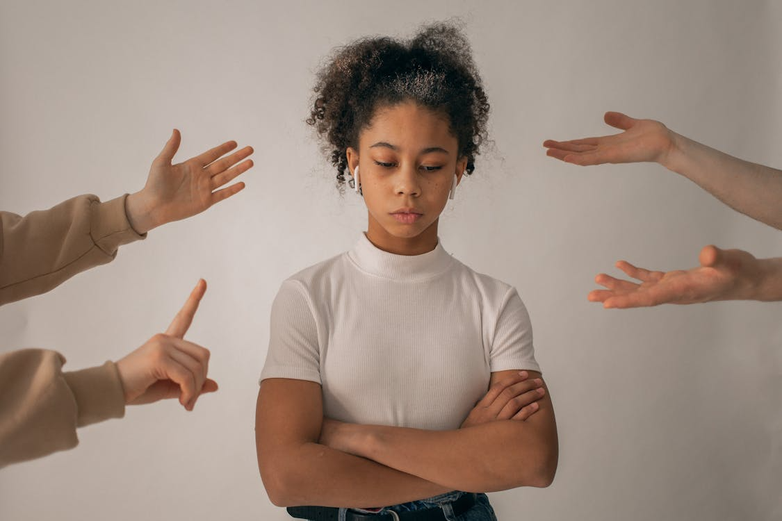 Sad girl with crossed arms wearing earbuds surrounded by hands pointing and gesturing at her.