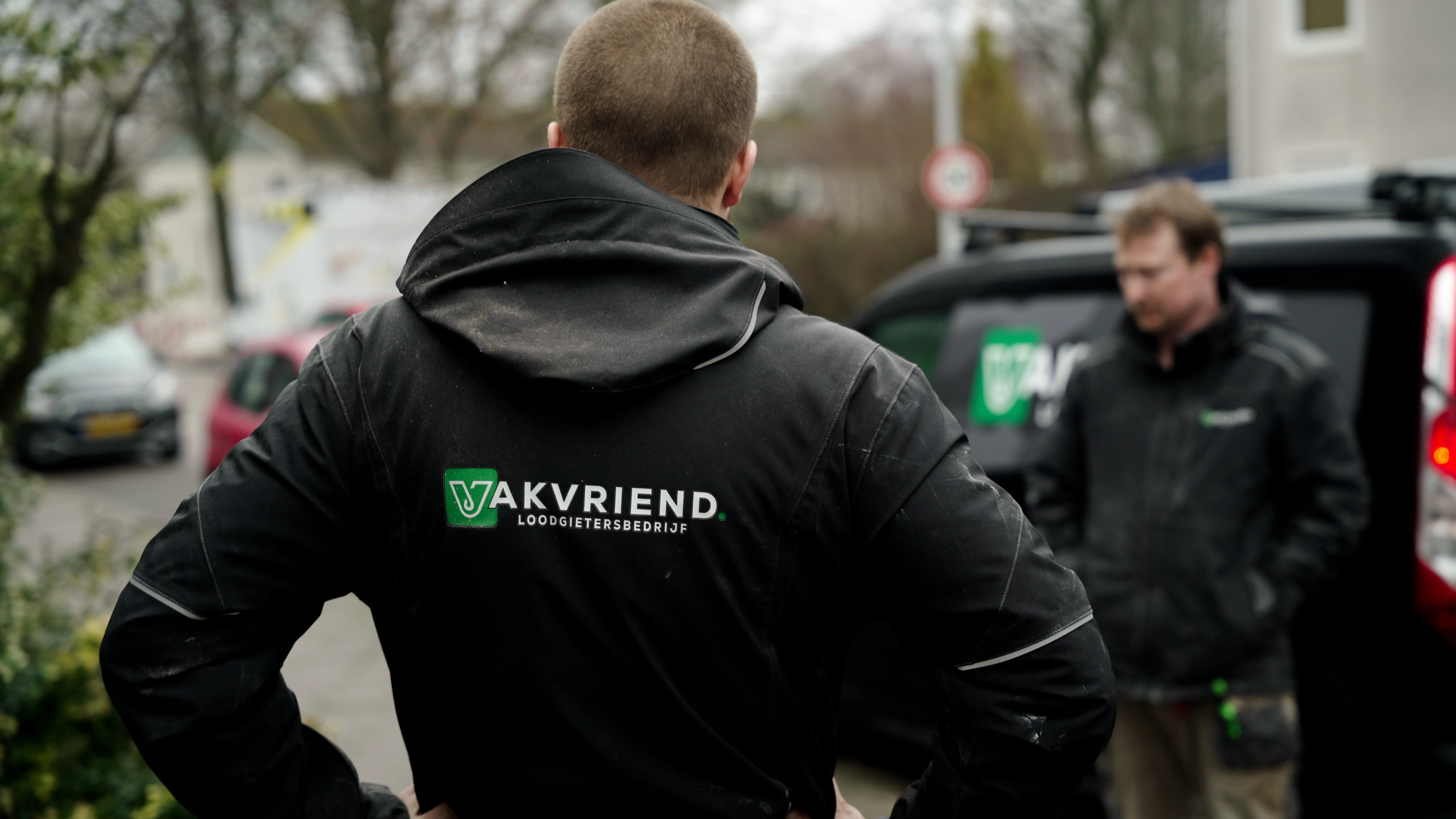 Een man gezien van achteren draagt een zwarte jas met het logo van loodgietersbedrijf Akvriend, terwijl een andere man in een vergelijkbare jas naast een bedrijfsbus staat.