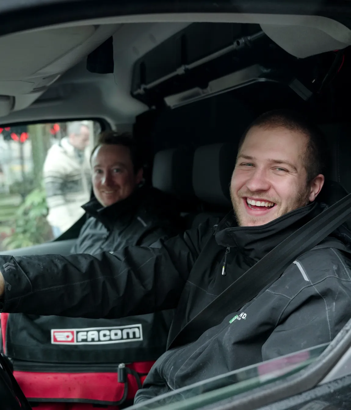 Twee mannen in zwarte jassen zitten in een voertuig, waarbij de voorste man lacht en een veiligheidsgordel draagt.