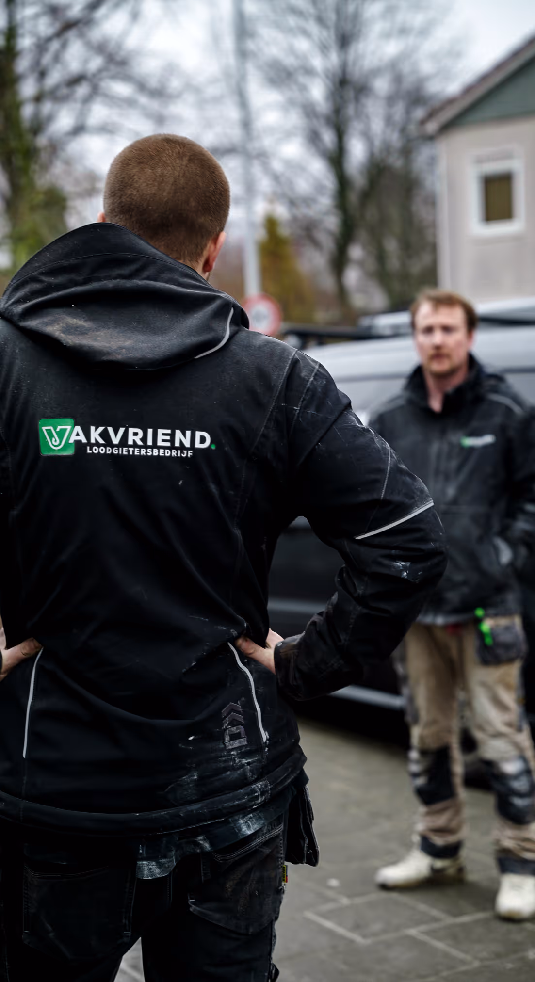 Twee mannen in zwarte jassen glimlachen terwijl ze in een voertuig zitten; de man rechts draagt een veiligheidsgordel.