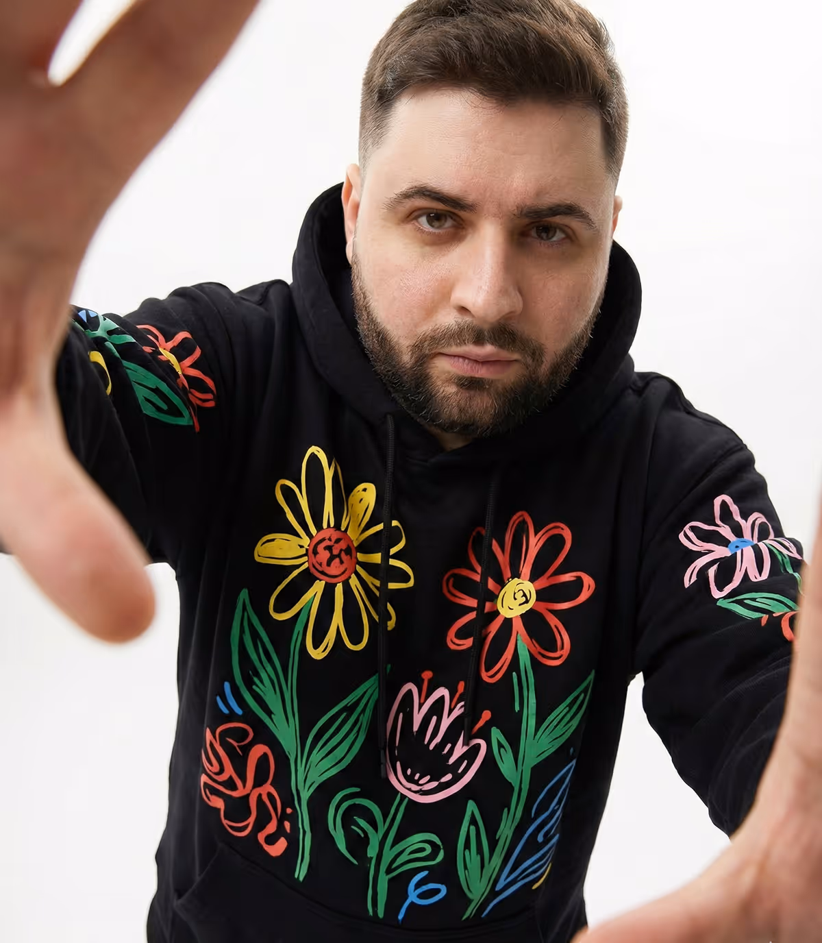 Man with beard wearing a black hoodie decorated with colorful flower drawings reaching towards the camera.