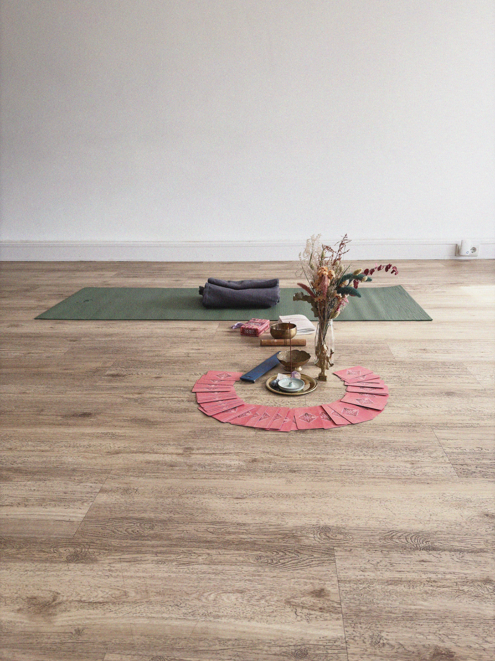 Grüne Yogamatte auf einem Holzboden mit einem gefalteten Handtuch, in einem Halbkreis angeordneten Tarotkarten, einem Räucherstäbchenhalter und einer Vase mit Trockenblumen in der Nähe.