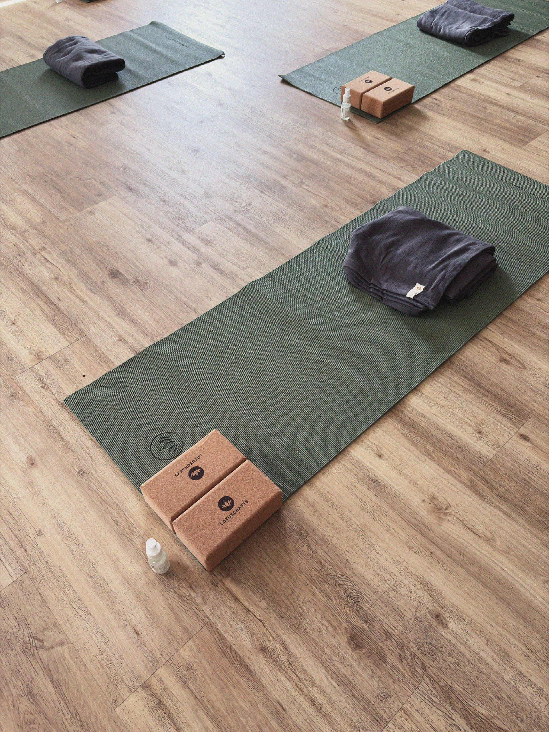 Drei grüne Yogamatten auf einem Holzboden, jeweils mit einem gefalteten grauen Handtuch, zwei Kork-Yogablöcken und einer kleinen Sprühflasche.