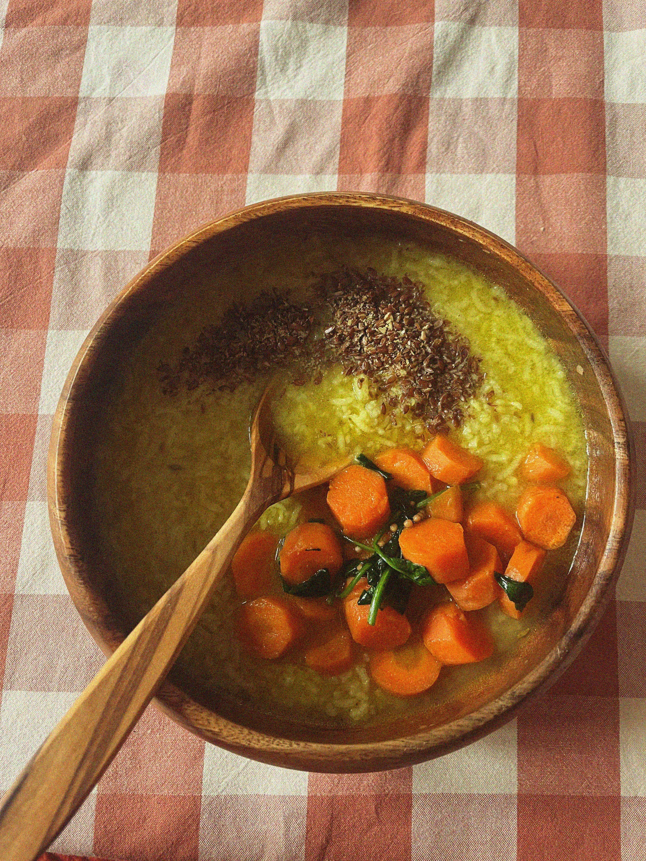 Holzschale mit einer gelben Reis-Gemüse-Suppe, die geschnittene Karotten, Blattgemüse und Leinsamen enthält, auf einem rot-weiß karierten Tuch.