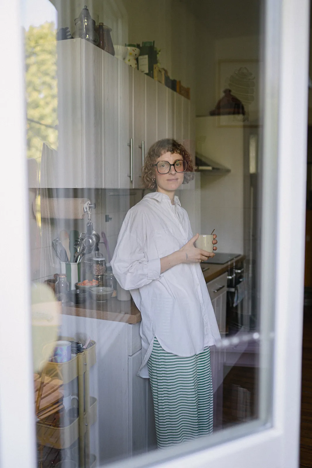 Junge Frau mit schulterlangem braunem Haar und schwarzem T-Shirt sitzt vor heller Leinwand mit Schatten und grüner Pflanze.