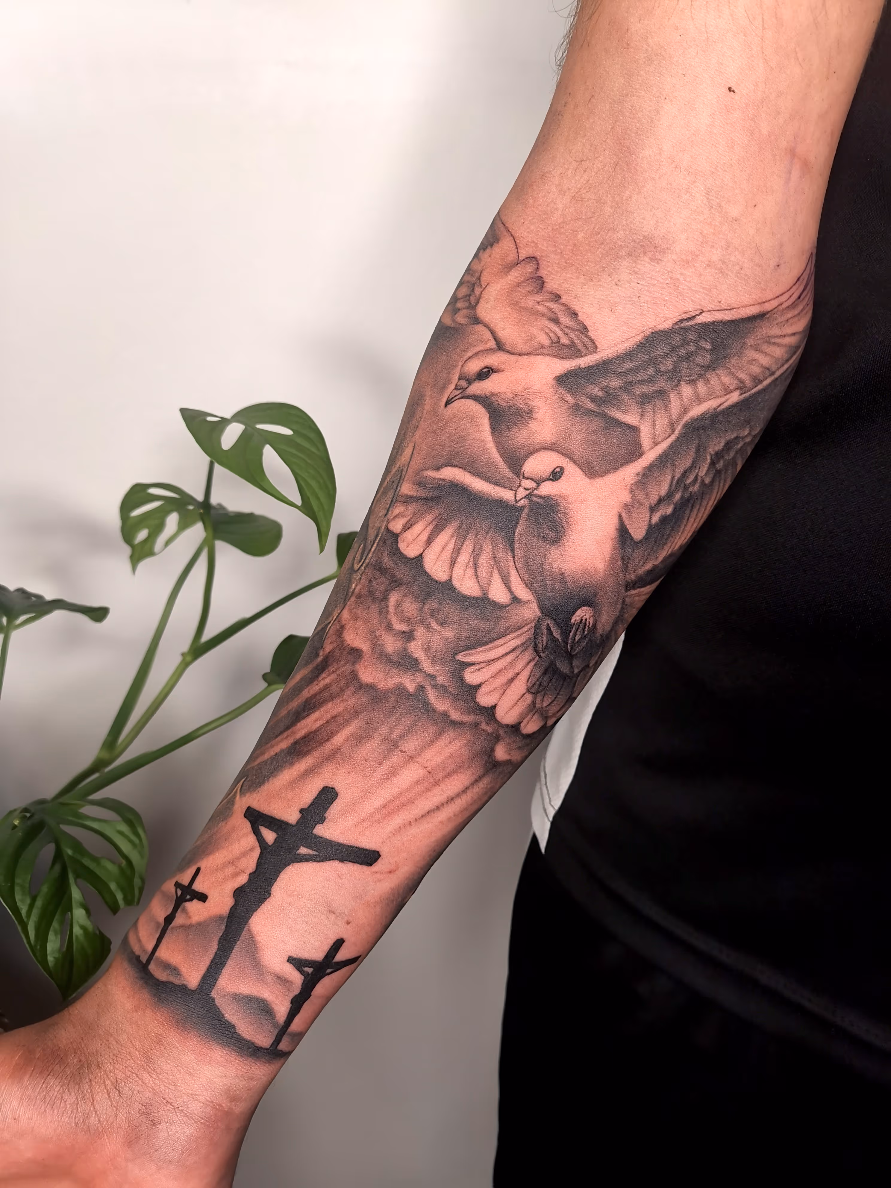 A forearm tattoo of two doves flying over a cloud with sun rays shining down on a mountains view with three crosses
