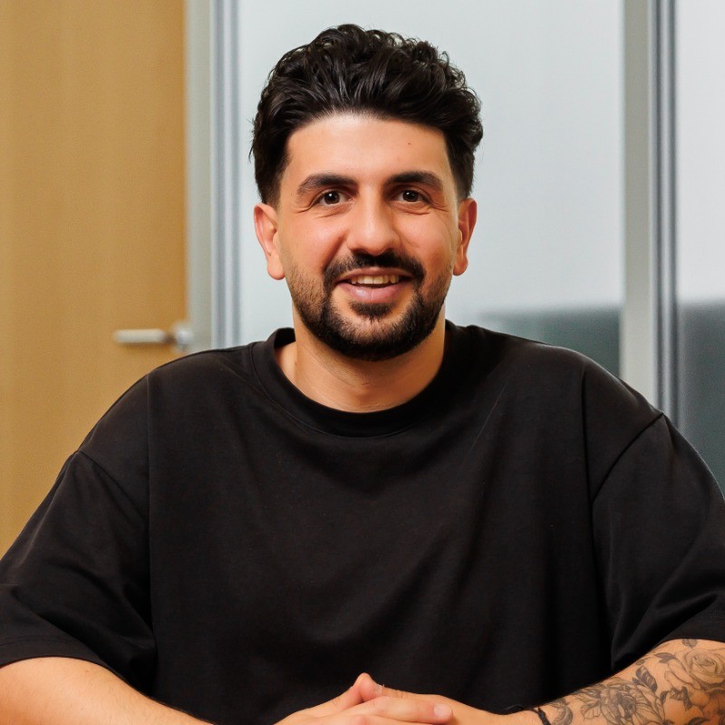 Lächelnder Mann mit dunklem Haar und Bart trägt ein schwarzes T-Shirt und zeigt Tätowierungen auf dem linken Unterarm.