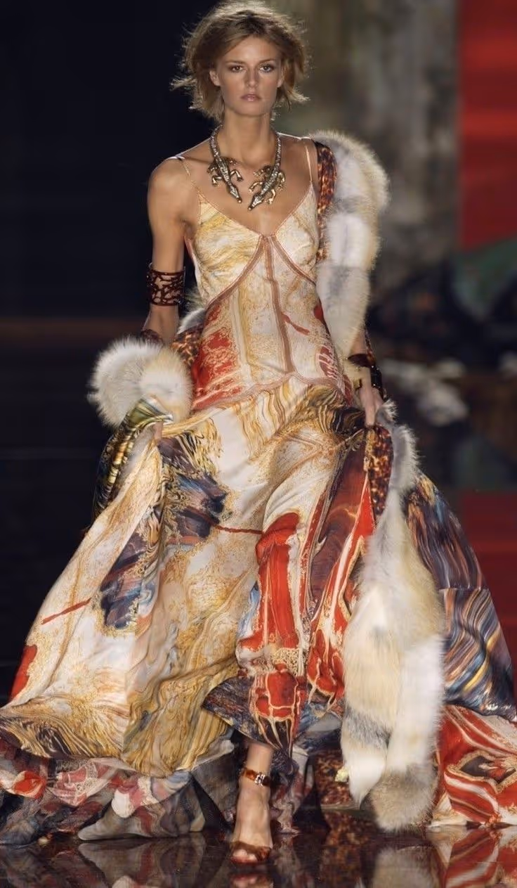 Mannequin défilant avec une robe longue fluide multicolore aux tons rouges, dorés et bleus, portant un accessoire de fourrure blanche et un collier doré imposant.