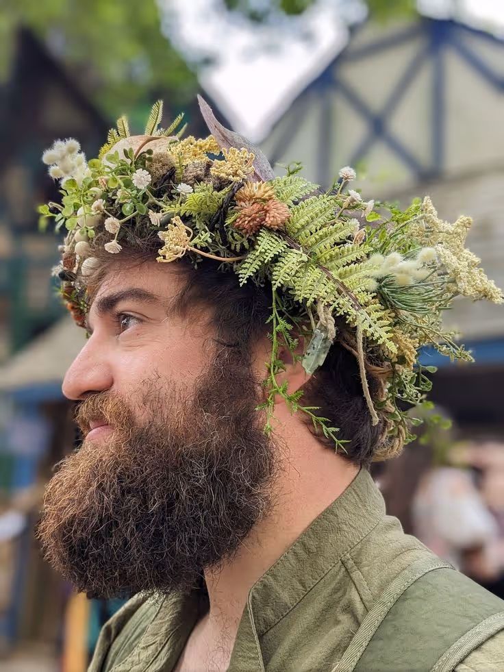 Homme avec une barbe généreuse portant une couronne de fleurs et de feuillages naturels, regardant vers la gauche.