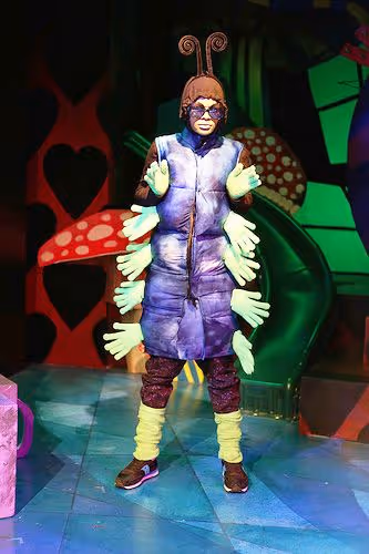 Personne déguisée en chenille avec costume violet, antennes, et plusieurs mains vertes attachées au costume sur scène colorée.