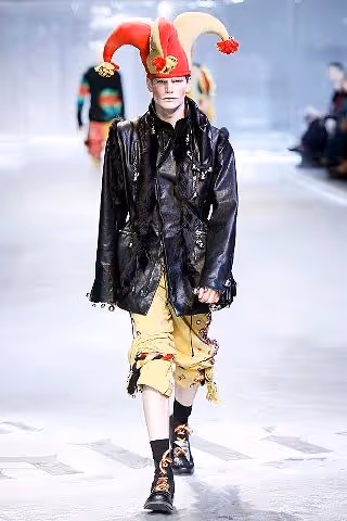 Un mannequin défile portant un chapeau de bouffon rouge et beige, une veste noire en cuir et un pantalon jaune avec des décorations, sur une piste défilé.