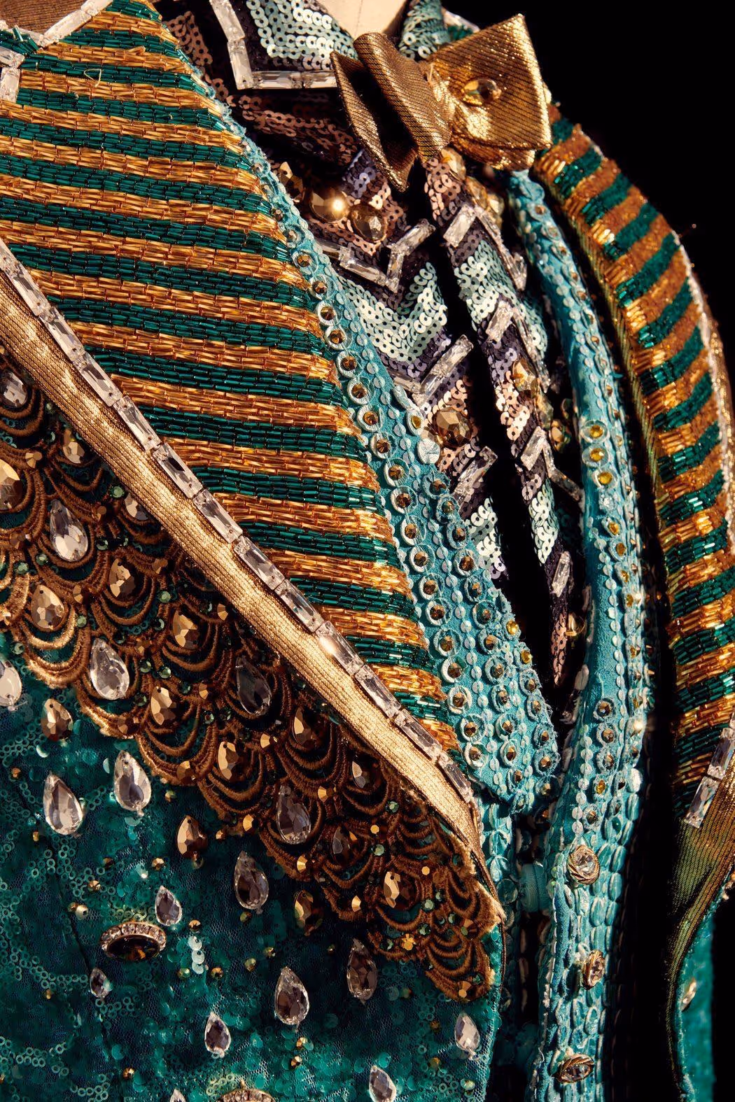 Détail d'un costume orné de perles dorées, sequins verts et or, pierres précieuses et motifs élaborés.