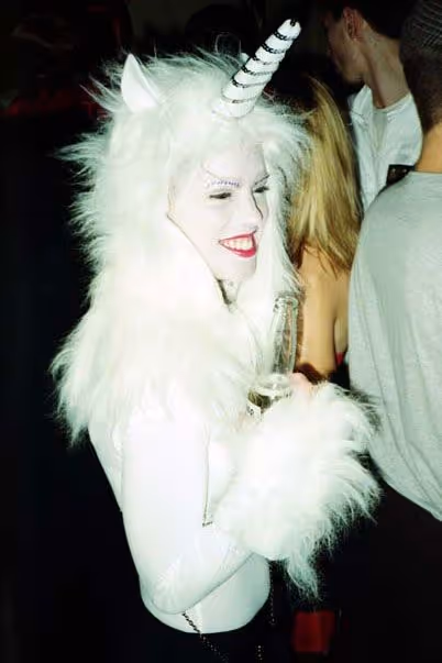 Personne déguisée en licorne blanche avec corne rayée, pelage doux, maquillage blanc et rouge à lèvres rouge, tenant une bouteille.