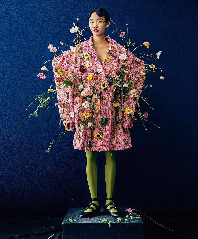 Modèle debout portant un manteau rose orné de nombreuses fleurs multicolores, des collants verts et des chaussures noires, sur un podium noir avec un fond bleu foncé.