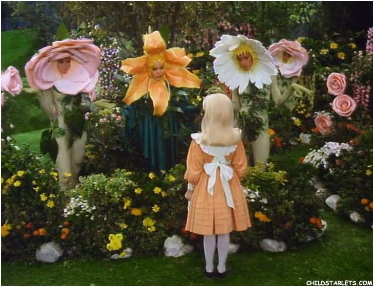 Une petite fille blonde en robe orange avec un tablier blanc fait face à trois personnes déguisées en fleurs colorées dans un jardin fleuri.