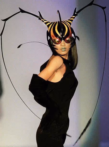 Personne portant un masque et un costume d'insecte avec de longues antennes noires et des détails jaunes et rouges, posant sur fond clair.