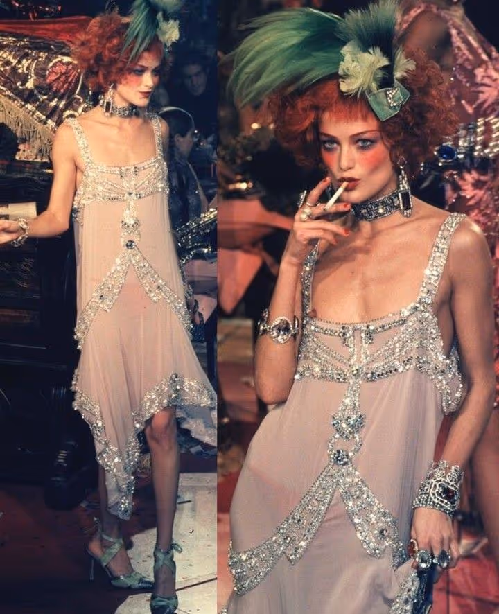 Femme aux cheveux roux bouclés et maquillage dramatique, portant une robe rose pâle ornée de sequins et un grand chapeau vert à plumes, fumant une cigarette lors d'un événement.