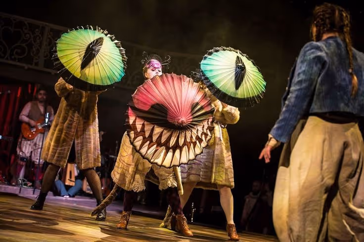 Trois artistes en costumes de scène tiennent des éventails colorés lors d'une performance théâtrale, avec un public en arrière-plan.