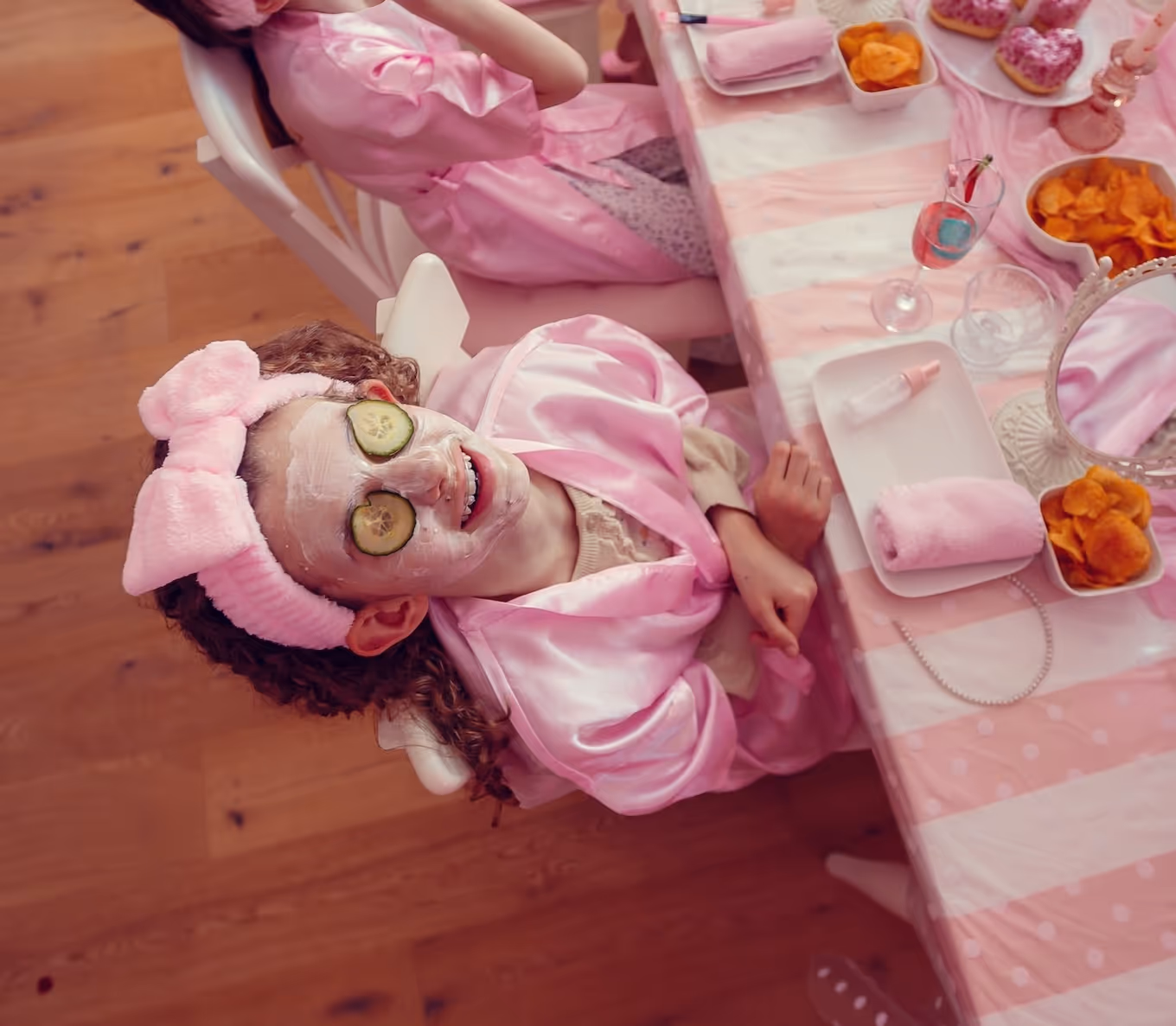 Kind lächelt mit Gesichtsmaske und Gurkenscheiben auf den Augen bei einem Pyjama-Party-Wellness-Setup.