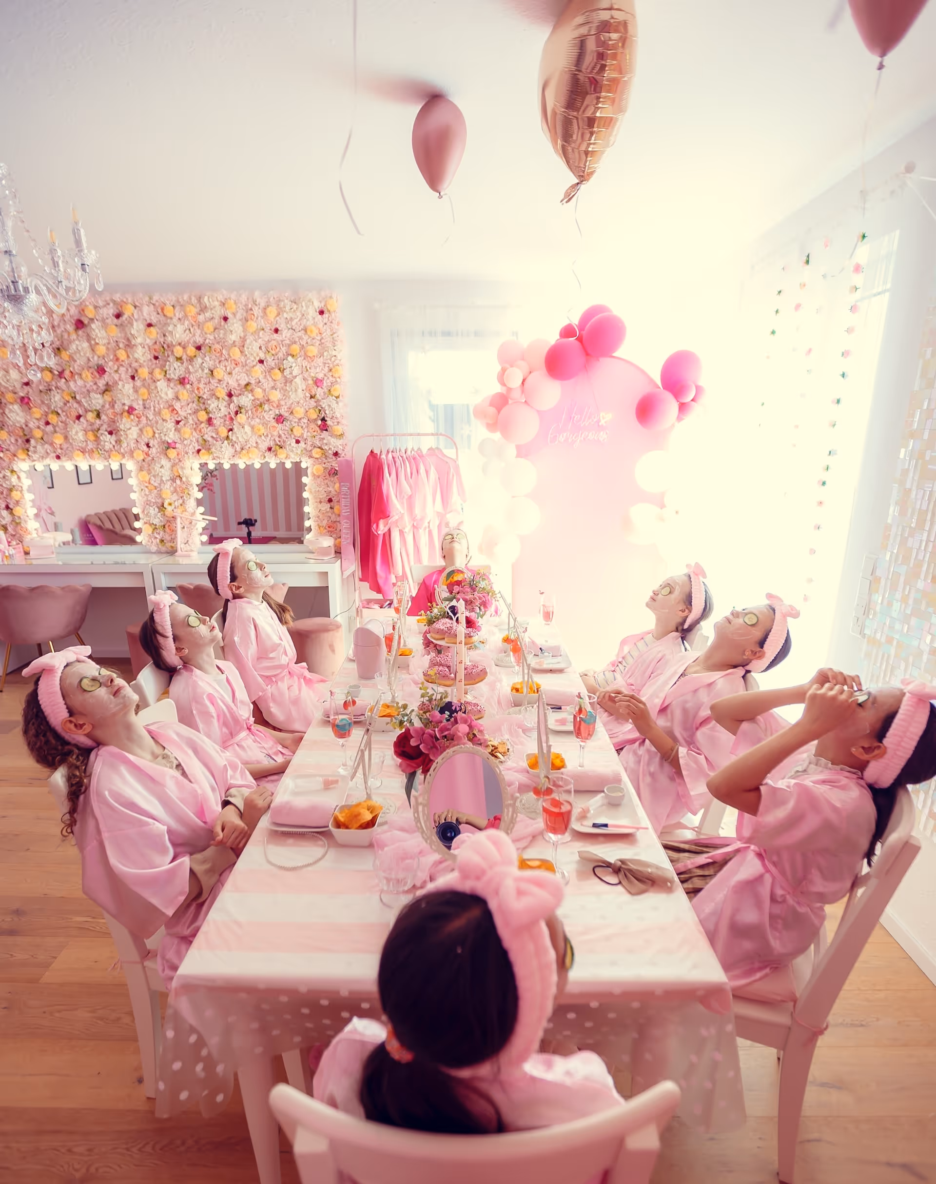 Acht Frauen bei einem Spa-Mädelsabend sitzen am Tisch mit Gesichtsmaske, Gurkenscheiben auf den Augen und rosa Bademänteln.