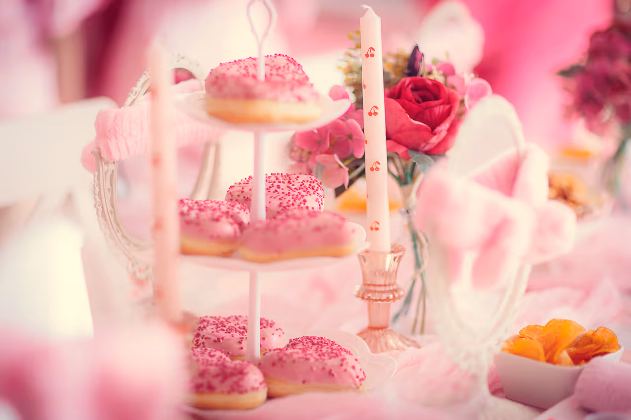 Dreistufiges Tablett mit rosafarben glasierten, herzförmigen Donuts, daneben eine Kerze mit Kirschen-Motiv und ein Blumenarrangement mit rosa Blüten.