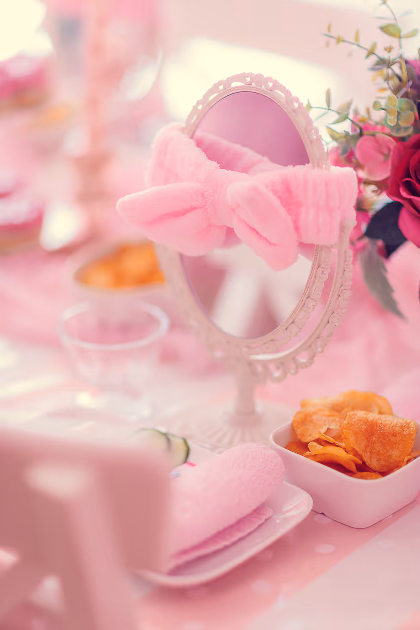 Runder Spiegel mit verziertem Rahmen, der ein rosa, plüschiges Stirnband mit Schleife trägt, neben einer Schüssel mit Chips und rosa Dekoration.