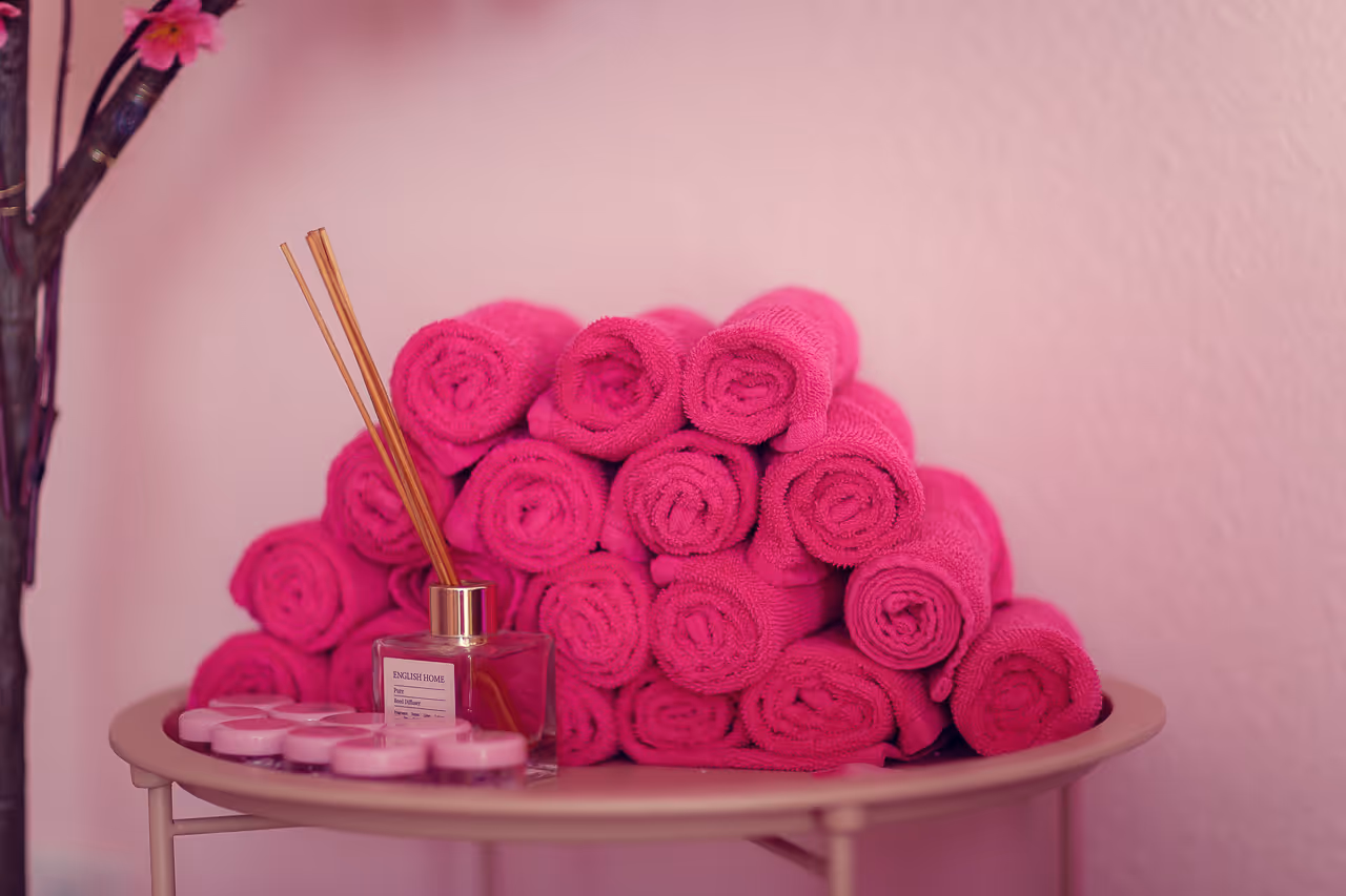 Gestapelte rosa Handtücher mit Duftstäbchen und kleinen pinken Behältern auf einem runden Tisch vor rosafarbener Wand.