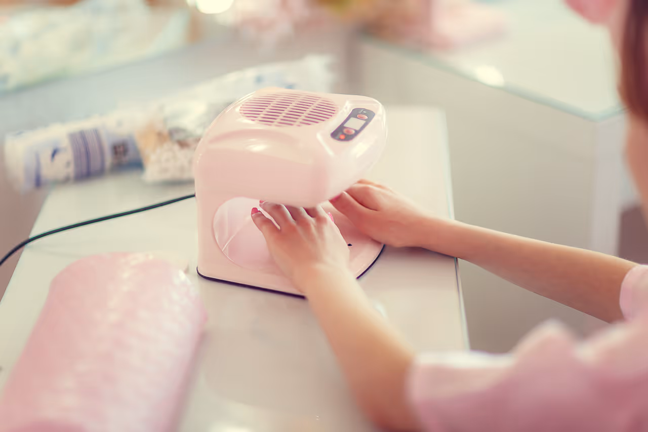 Person legt Hände unter eine rosa Nageltrocknermaschine auf einem weißen Tisch.