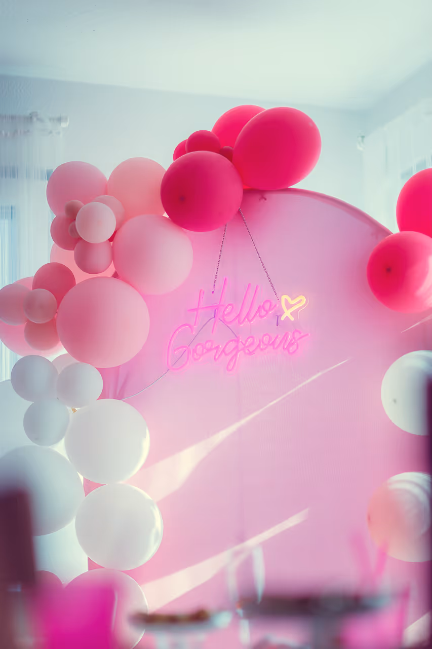 Runder rosafarbener Hintergrund mit rosa, pinken und weißen Luftballons und einem leuchtenden Schild mit der Aufschrift ‚Hello Gorgeous‘.