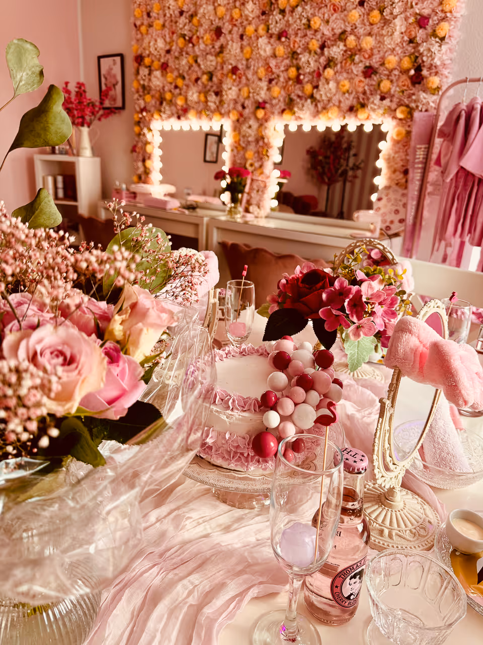 Festlich dekorierter Tisch mit pink-weißer Torte, Blumensträußen, Sektgläsern und rosafarbenem Saft in einem rosa dekorierten Raum mit beleuchtetem Spiegelfeld.