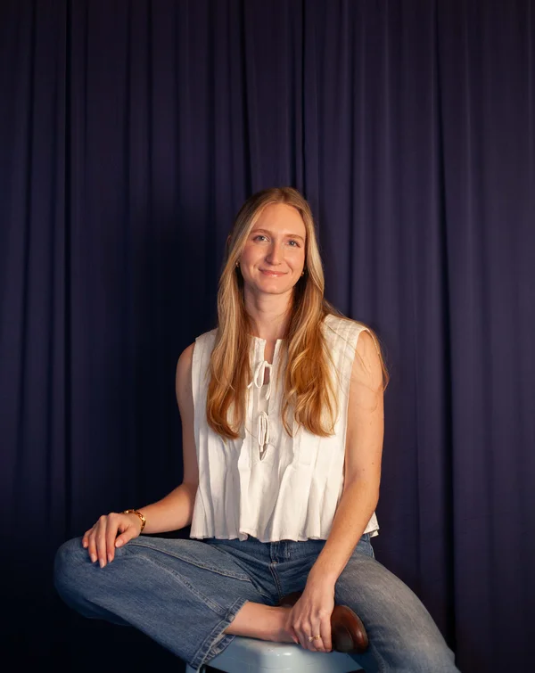 Smiling young woman with long blonde hair wearing a white sleeveless top and blue jeans, seated cross-legged on a stool against a dark curtain background.