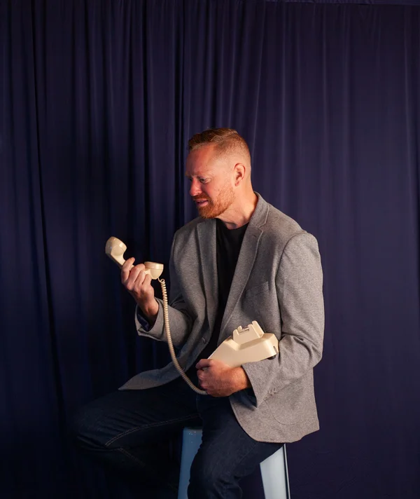 Man with red hair and beard in gray blazer sitting and looking at a beige rotary phone handset.