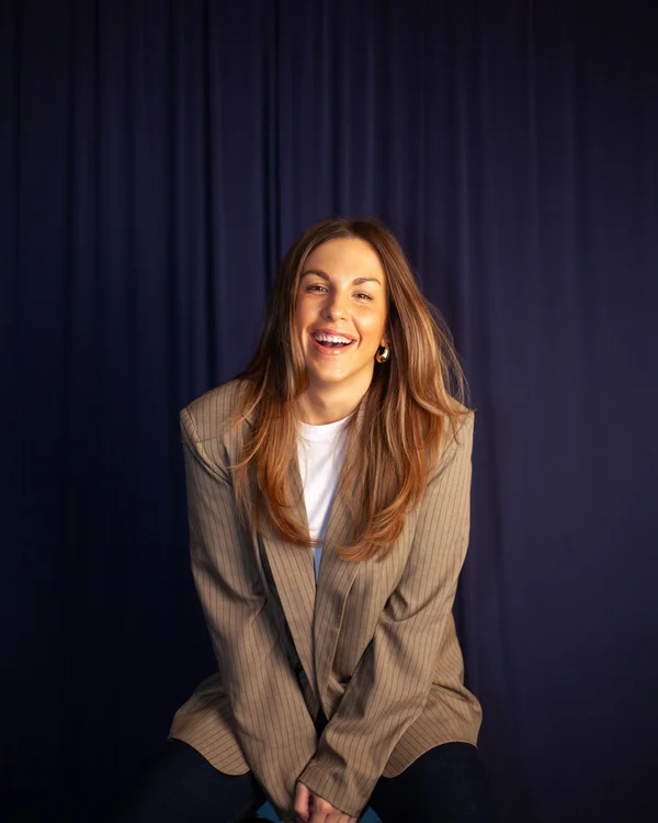 Smiling woman with long brown hair wearing a pinstripe blazer and white shirt against a dark blue backdrop.