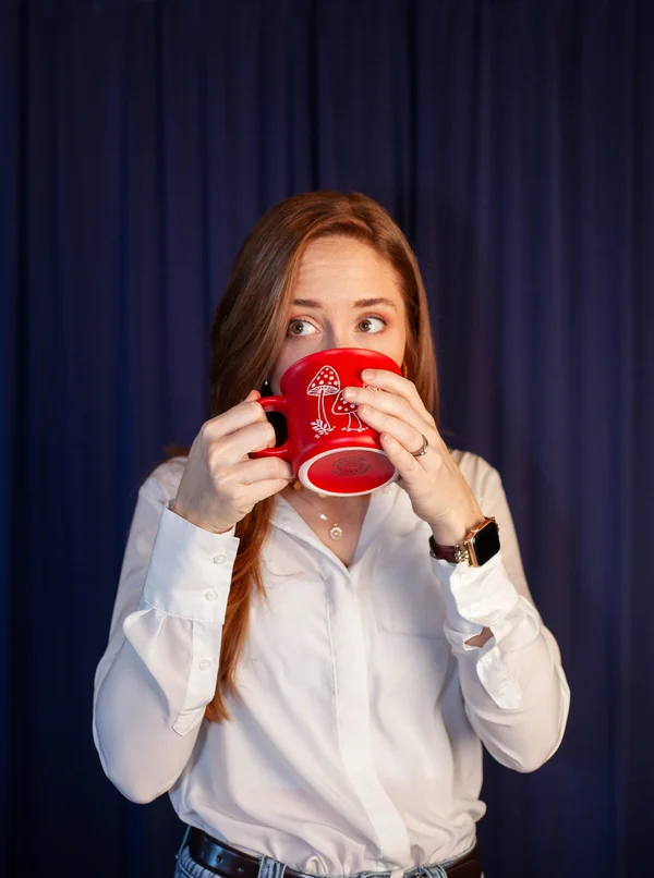 Woman with light brown hair wearing a white blouse drinks from a red mug with mushroom illustrations against a dark blue curtain background.