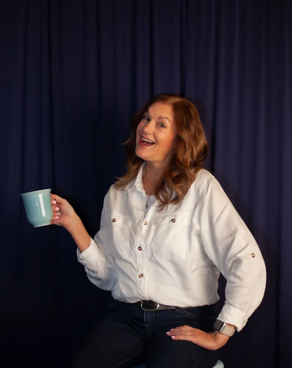 Smiling woman with wavy brown hair wearing a white button-up shirt and dark jeans, holding a light blue mug against a dark curtain background.